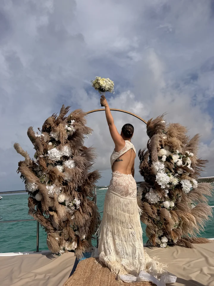 Novia levantando su bouquet junto a un arco con pampas en un catamarán de Punta Cana