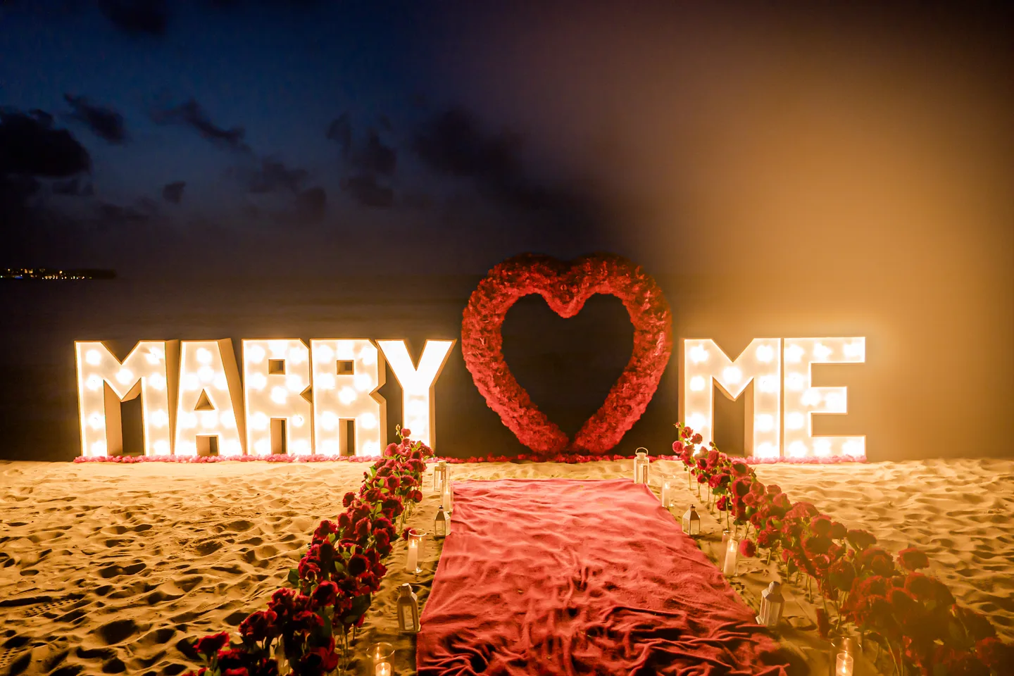 Montaje de propuesta al aire libre con rosas rojas y letras Marry Me iluminadas